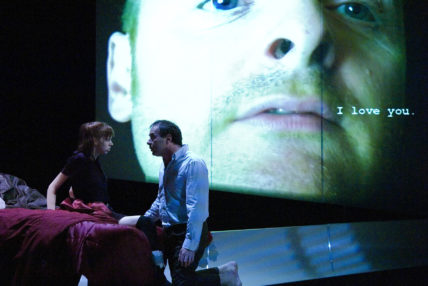 Image of two cast members kneeling, with large projected face behind.