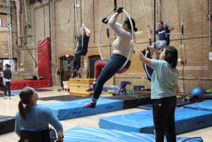 A group image with a woman on a trapeze and two men in the background on the bar