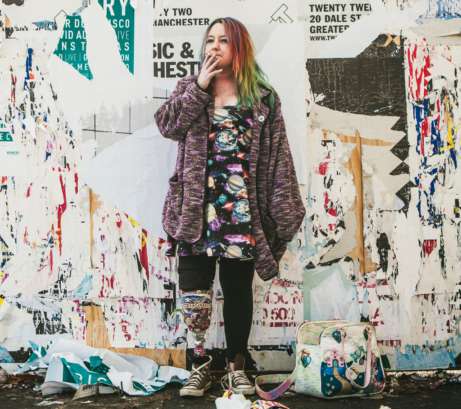 A photo of Jackie Hagan, standing in front of a wall of old posters, smoking a cigarette.