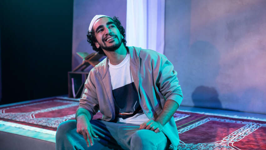 A young Asian man with black curly hair and short facial hair smiling, sits on an ornate red and white rug, looking off to one side, to something in his distance. He wears a topi hat, a beige open cardigan and white t-shirt underneath. A white curtain hangs behind him.