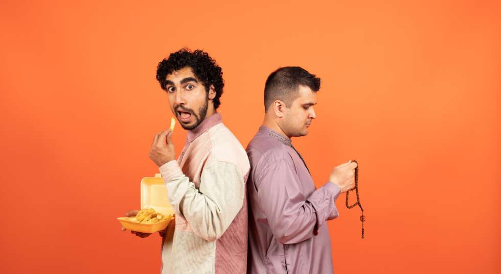A photo showing two men standing back to back, wearing traditional Muslim dress. The man on the left eats chips, with his mouth open, looking to the camera, while the man on the right looks down, holding prayer beads. They stand against a bright orange background.
