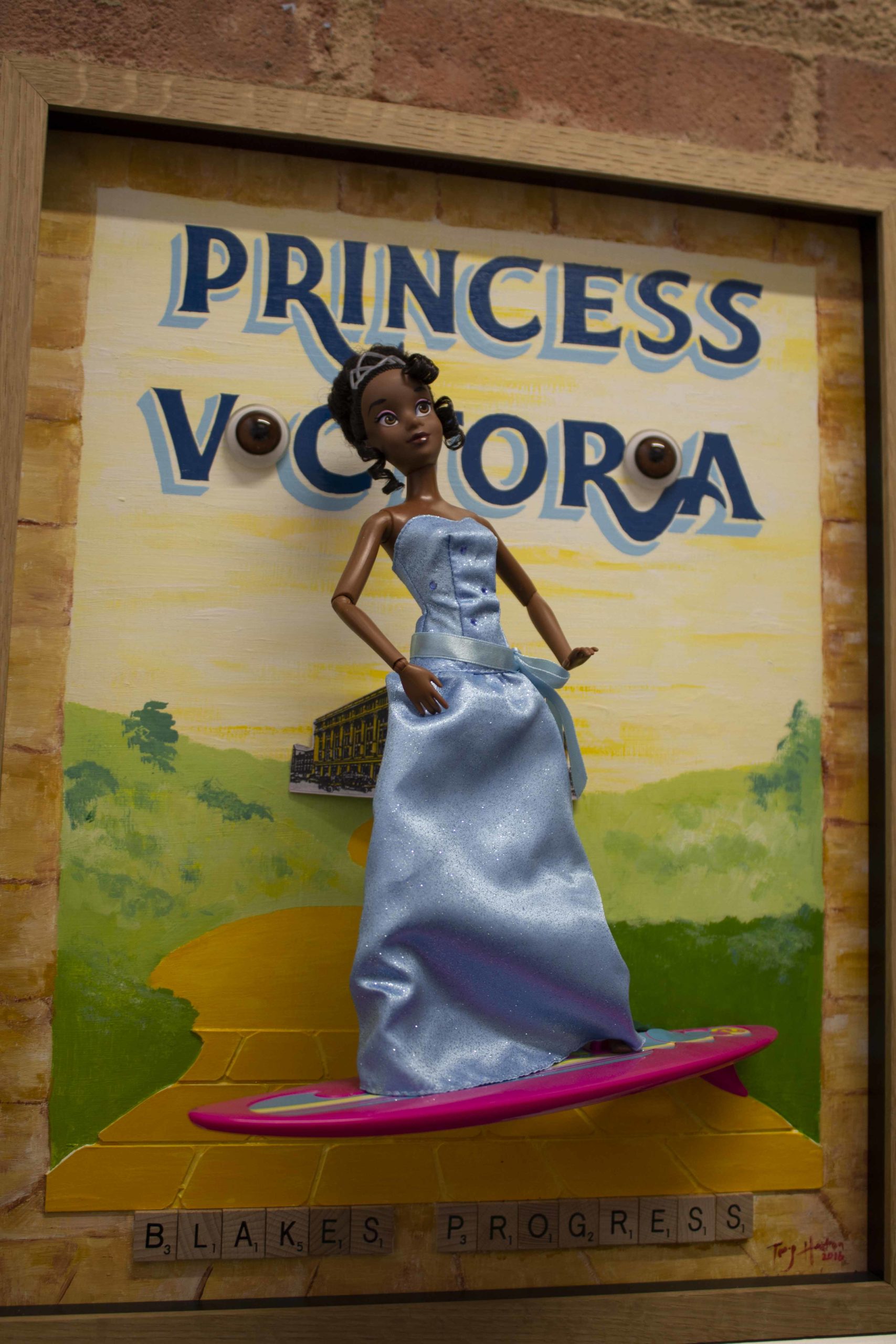 The background of the piece is a pale yellow sky, above a green landscape, either side of a yellow brick road. At the top of the page in blue letters are painted the words ‘Princess Victoria’. The letters I in Victoria have been replaced with three dimensional brown glass eyes. Protruding from the canvas is a black, Barbie-style doll in a pale blue evening dress and tiara, standing on a hot pink surf board.