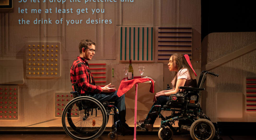 Two young white wheelchair users, male and female, sit opposite each other at a table with a red table cloth, on a date. Text projected behind them reads "So let's drop the pretence and let me at least get you the drink of your desires".