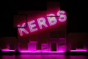 Text reading KERBS projected in bright pink neon onto a wall, on an otherwise dark set.