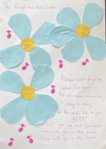 This piece is by Kathryn Greenwood. It is a mixture of an image and a poem. On the left hand side of a white background are large blue flowers made from blue paper with a yellow orange paper centre. There are small pink music notes surrounding the flowers. The piece has a title: The Forget me Not Choir, it is accompanied by the following poem: Please don't forget me when I'm gone, put on some music or sing a song. Let the music talk to your heart lift you up and feel great. I am with you in the music, I am with you in the heart. The artist chose to make this piece as The Forget me Not Choir is incredibly important to her. The Forget me Not Choir, is a choir that helps those the terminal illnesses socialise, sing and enjoy life.