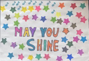 This images by L Humphreys. In the middle of a white background are the words “may you shine” written in capital letters. Each letter is a different colour. Surrounding the words are a mixture of music notes and stars in many different colours. The artist chose to put stars in many different colours to represent all the different people within a community and although we may have a colour that is our favourite or represents a certain aspect of our personality we're all stars and we should shine together.