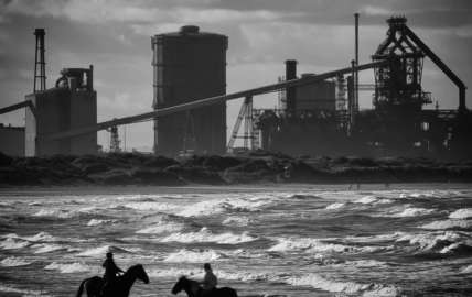 A black and white photo of two people riding two horses on the beach with construction/factory work in the background