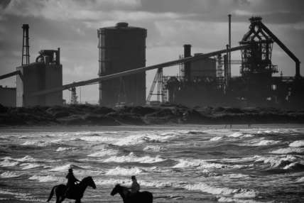 A black and white photo of two people riding two horses on the beach with construction/factory work in the background