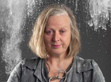 Poster image of Jenny Sealey. A white woman wearing a dark grey dress with a curly silver necklace and pink lipstick. Jenny looks to camera with a bewildered expression. There is a cloud of flour above her head about to fall.