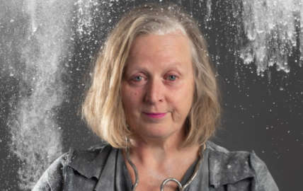 Poster image of Jenny Sealey. A white woman wearing a dark grey dress with a curly silver necklace and pink lipstick. Jenny looks to camera with a bewildered expression. There is a cloud of flour above her head about to fall.