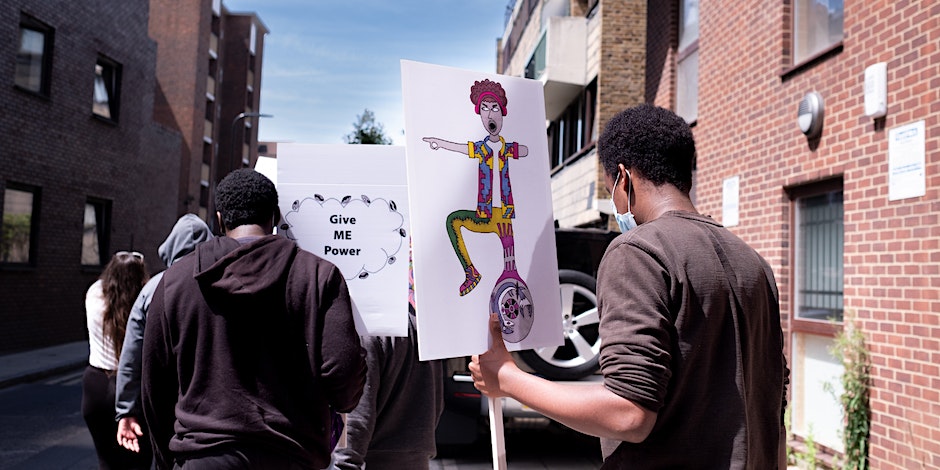 The back of two young black men holding a protest sign which reads 'give me power'